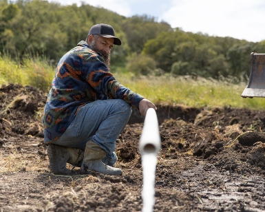 A farmer watches water come out of a plastic pipe and onto some dirt.
