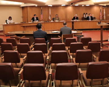 A view of the March 24 Board of Supervisors meeting ,showing mostly empty seats in the foreground and the Board members sitting further apart from usual at the dais.