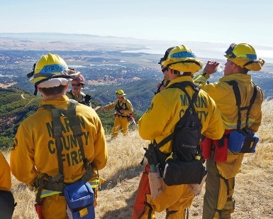Several firefighter trainees in their 20s, wearing fireproof turnout gear, stand atop a hill overlooking San Francisco Bay during a pause in training.