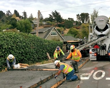 Construction workers flow wet concrete out of a cement mixer truck and onto a sidewalk.