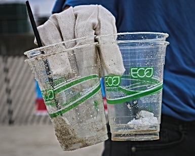 A closeup view of a person's gloved hands holding two plastic cups that are not recyclable. 