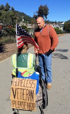 Veterans Service Officer Sean Stephens stands next to a man in a wheelchair who has a sign saying 'Homeless Veteran, Please Help'