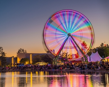 An illuminated ferris wheel spins at the County Fair, with the reflection of the lagoon in the foreground