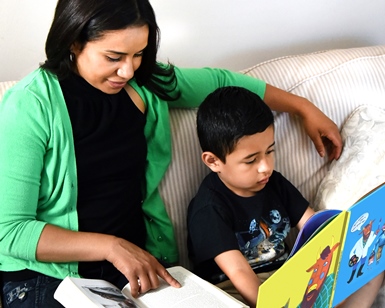 A woman reads a book with her arm around a young boy as he reads his own book.