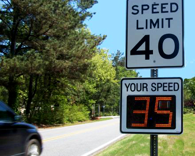 A roadside speed limit sign shows an additional attachment on the pole with an electronic display that shows the passing driver's radar speed.