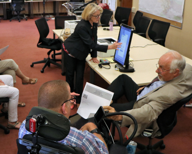 A man in a wheelchair receives a demonstration on how accessible voting equipment works from the female registrar of voters, who is standing and pointing to a computer screen.
