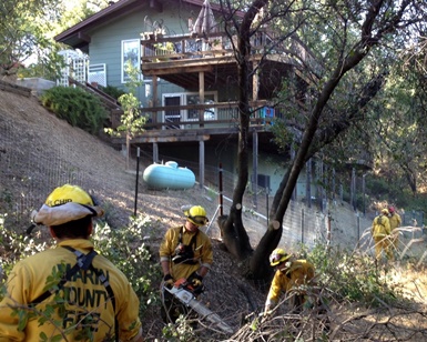 Three firefighters prepare debris for a pileburning session with a home in the background.