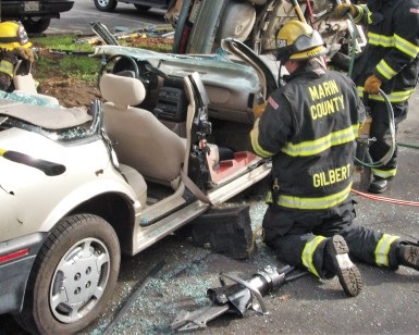 Three firefighters work on a mangled car during an extrication drill as a "jaws of life" device sits on the pavement in the foreground.