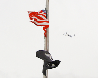 A view of a flagpole with an American flag at half staff and a POW MIA flag below it, with five antique airplanes flying in the distance.