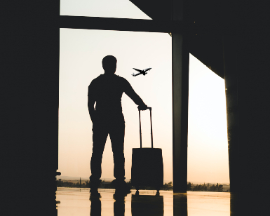 A photo shows a silhouette of a male traveler with a suitcase standing in an airport lounge and looking through a window at an airplane taking off.