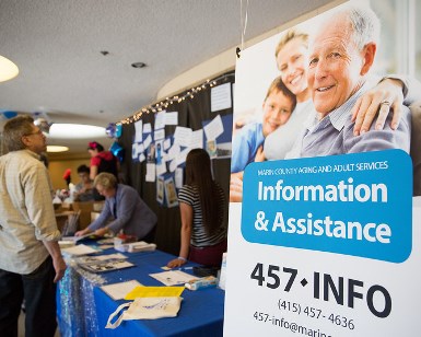 On the right is a 5-foot-tall sign promoting the Information & Assistance phone number 415-457-INFO. On the left are people mingling at an information table at senior fair.
