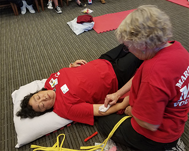 Two female volunteers from Marin Medical Reserve Corps participate in a 2019 disaster drill. One is lying on the ground as a medical patient, the other is administering first aid to the person lying on the ground. 