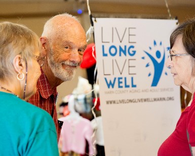 An older man smiles at the camera next to a sign that says Live Long Live Well.
