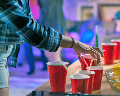 A closeup view of a party scene shows the hands of a young person touching plastic cups presumably with alcoholic beverages in them.
