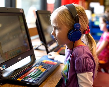 A girl of about 6 years old wears headphones and looks at library computer screen