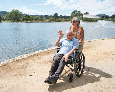 A woman pushed a wheelchair occupied by an elderly man who is waving as they roll on an unpaved pathway around the Civic Center Lagoon in San Rafael.