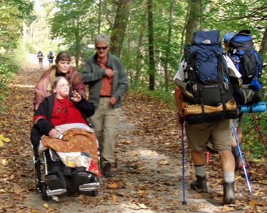A woman in a wheelchair is pushed by another woman on an unpaved trail as other hikers walk by.