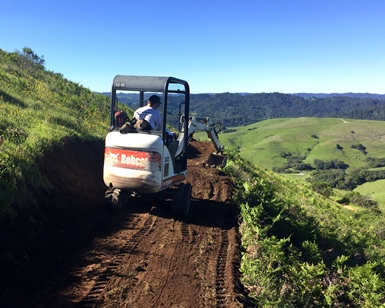 A man in a small Bobcat tractor works creating on a dirt trail with rolling hills in the background.