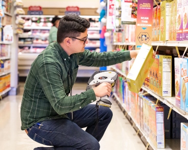 A male weights and measures inspector kneels down in a grocery store aisle to check the price of an item with a hand-held device.