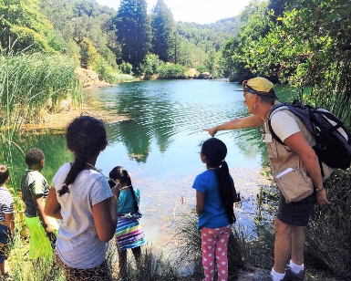 A man points to water in a lake as young kids look to the water.