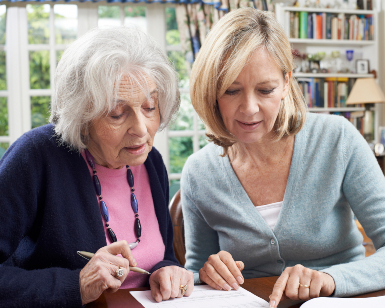 An older woman on the left examines documents on a table as a younger woman assists her.