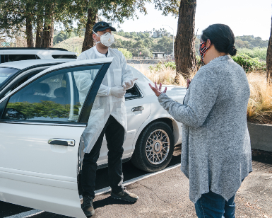 A woman (right) gives directions to a man (left) standing outside of a vehicle after he has put on protective smock, gloves and a mask in preparation for driving somebody to a COVID-19 testing location.