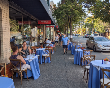 A view of a downtown business district with diners sitting at a sidewalk table, a man walking on the sidewalk, and a car driving by.