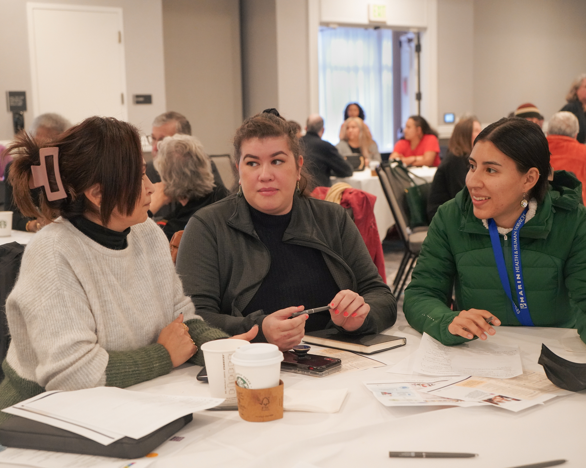 Three women speak about participatory budgeting while sitting at table in a large room of people.