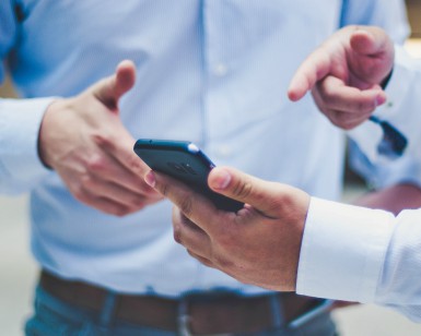 A tight shot of the hands of two men, with one man holding a cell phone and pointing to it with his other hand.