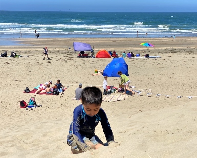 A young boy plays in the sand at a local beach with other people sunbathing and playing in the background
