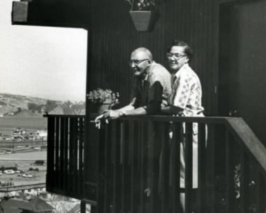 An older man and woman stand on a home's deck with the bay and hills in the background.
