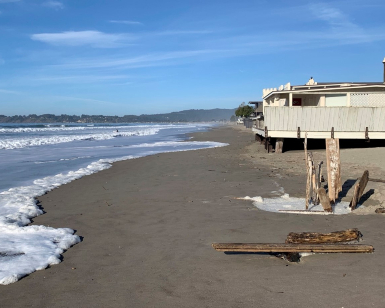A view from the sand at Stinson Beach with a home on short stilts to the right and the ocean to the left.