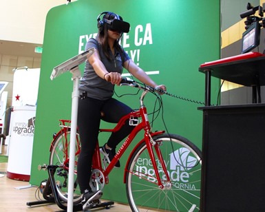 A woman rides a stationary bike while wearing a headset and googles as she demonstrates the virtual bike tour.