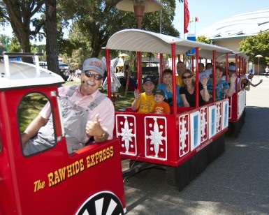 A small trolley carries fairgoers around the fairgrounds as people wave and the engineer gives a thumbs-up sign.
