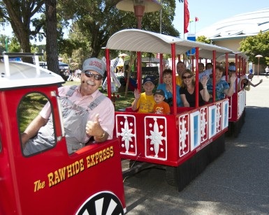 A small trolley carries fairgoers around the fairgrounds as people wave and the engineer gives a thumbs-up sign.