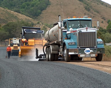 A County crew works on paving a road, with a big truck and tractor on the right and freshly laid gravel on the left.