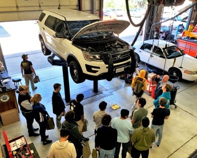About a dozen teen students look at the underside of an SUV up on a hydraulic lift inside the County Garage as a mechanic describes the fleet maintenance program.