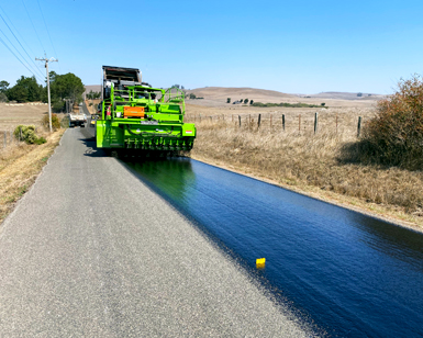 A large piece of heavy machinery lays down fresh black coating on a roadway that is being rehabilitated.