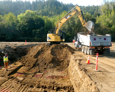 A large road excavator is shown dumping earth into a large dump truck during a flood control project.