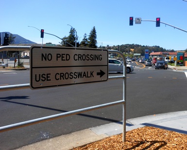 A sign at a busy intersection says "No Ped Crossing, Use Crosswalk" with an arrow pointing to the right.