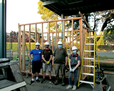 Four young students wearing helmets and face coverings stand in front of a wooden shed frame they made for the Department of Public Works. 