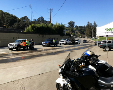 A California Highway Patrol motorcycle is on the right and several trucks with untarped loads are lined up in the background as the CHP officer talks to a truck driver.