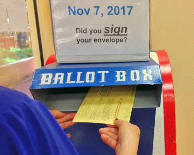 A closeup of someone's hand sliding a signed ballot envelope into a ballot box at the Election's Office.