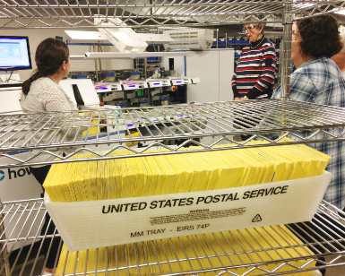 A view of the elections department equipment room, with a tray of ballot envelopes on a shelf in the foreground and three workers talking in the background.