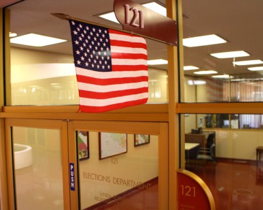 An exterior view of the Elections Department showing an American flag hanging out in front of the glass doors.