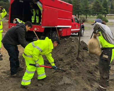 Three men wearing rain slickers shovel sand into bags during a storm response.
