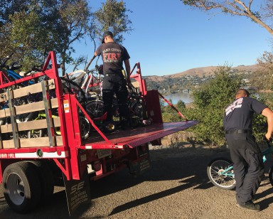 One male firefighter stands in a truck loaded with bikes, and a second male firefighter guides a bike away from the truck.