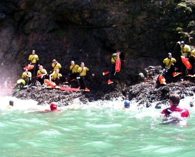 About 10 firefighters wearing wetsuits on a rocky beach and rough ocean water in the foreground during a training session.