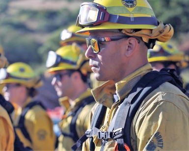 An African American firefighting recruit stands and listens to instructions with other firefighters during a training session.