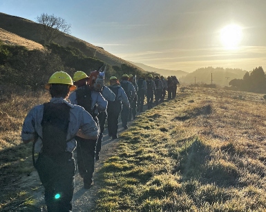 A view of a line of young firefighters wearing their gear and walking on a trail away from the camera toward a work assignment. 